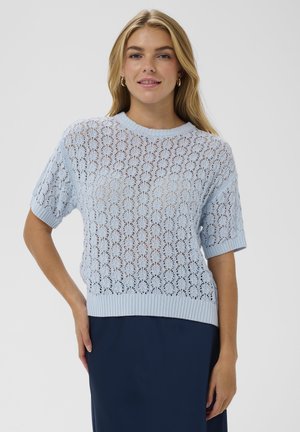 Femme portant un haut en maille de dentelle à manches courtes bleu clair avec un motif floral et une jupe bleu foncé, debout avec une main sur la hanche.