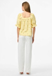 Blusa gialla con motivo floreale, maniche a sbuffo e scollatura quadrata, abbinata a pantaloni bianchi a gamba dritta, su uno sfondo chiaro.