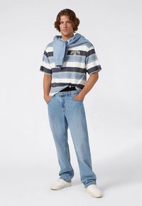 Camisa de manga corta a rayas en azul, gris y blanco, combinada con jeans azules. Una sudadera con capucha azul claro está atada alrededor del cuello. Zapatillas blancas.