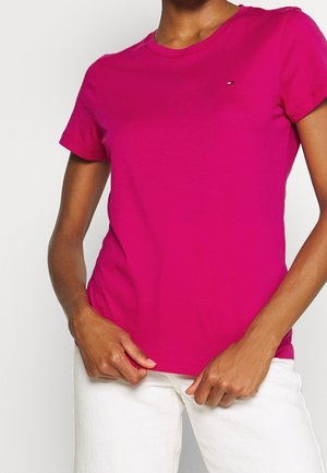 Osoba ubrana w jaskraworóżowy t-shirt z krótkim rękawem z małym logo na klatce piersiowej i białe spodnie na jednolitym tle.