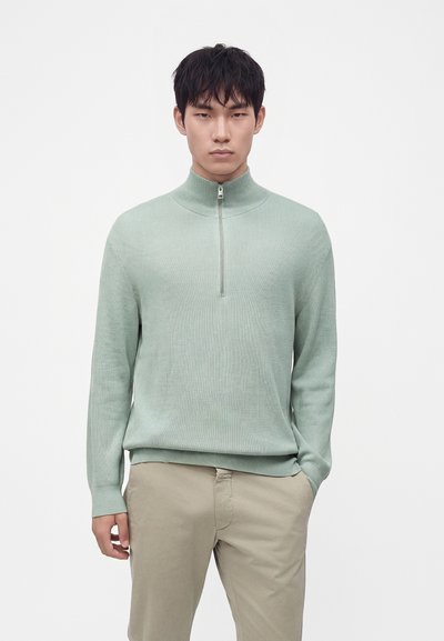 Homme portant un pull vert clair à demi-fermeture éclair et un pantalon beige, debout avec une main dans la poche devant un fond uni.