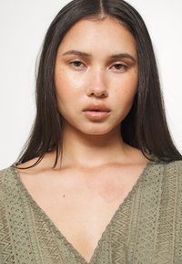 Olivgrön topp med V-urringning, med texturerade mönster och scallopade kanter. Slät tyg med synliga hudtoner under.
