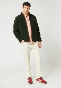 Giacca in pile verde con dettagli in zip rossi, indossata sopra una camicia a quadri leggeri, abbinata a pantaloni crema e scarpe da ginnastica rosse.