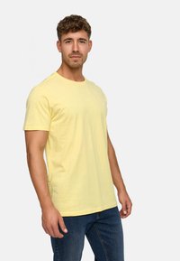 T-shirt en coton jaune �à col rond et manches courtes, avec un logo discret sur la poitrine et une coupe décontractée. Porté avec un jean bleu.