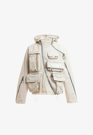 Chaqueta beige con capucha con múltiples bolsillos utilitarios con cremallera y botones a presión, cremallera delantera asimétrica y detalles de tiradores en forma de anilla plateada.