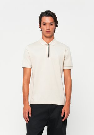 Polo à manches courtes en coton beige clair, avec un col zippé et une rayure contrastante, au design ajusté. Pantalon noir.