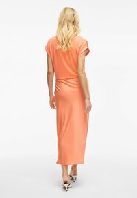 Robe maxi de couleur pêche avec des manches courtes, texture lisse et silhouette ajustée, associée à des mules à talons blancs.