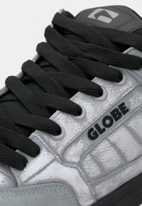 Close-up of a silver Globe sneaker with black laces and textured leather detailing on the side panel.