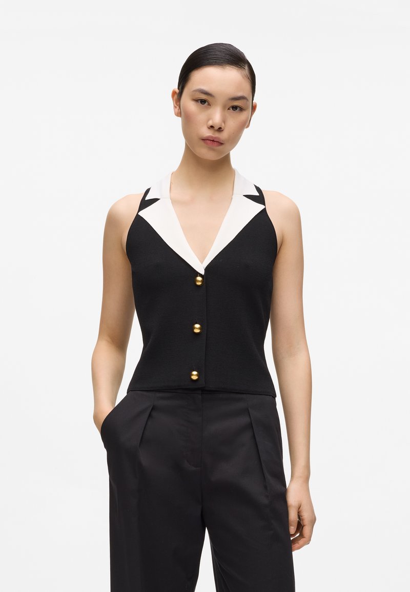 Γυναίκα που φοράει αμάνικο μαύρο γιλέκο με λευκό γιακά και χρυσά κουμπιά, συνδυασμένο με ψηλόμεσο μαύρο παντελόνι, στέκεται με το ένα χέρι στην τσέπη.