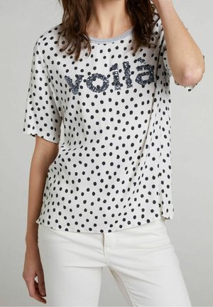 Donna che indossa una camicia bianca a pois neri con la scritta "voilà" in paillettes nere abbinata a pantaloni bianchi su uno sfondo semplice.