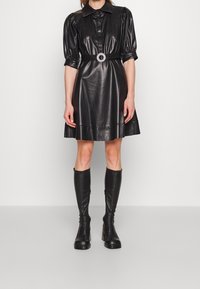 Robe en cuir noir avec des manches bouffantes, un devant à boutons, et une ceinture avec une boucle ronde. Des bottes en cuir noir montantes jusqu'aux genoux complètent l'ensemble.