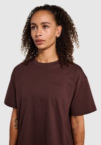 Bruine katoenen t-shirt met korte mouwen, voorzien van een ronde halslijn en een geborduurd "HALO" logo op de linkerborst. Gladde textuur.