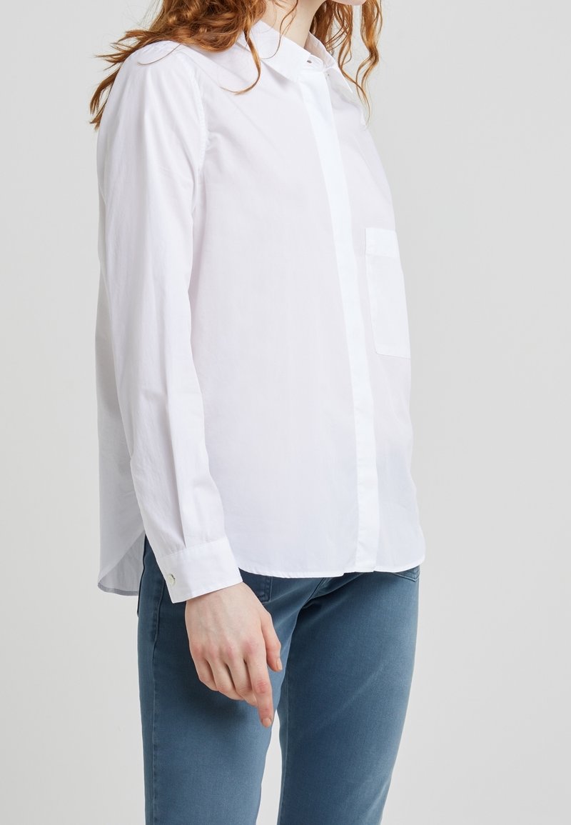 Camicia bianca a maniche lunghe con colletto abbottonato, taschino singolo sul petto e orlo curvo. Realizzata in un tessuto liscio. Abbinata a jeans blu.
