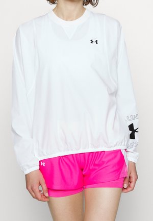 Personne portant un haut blanc ample à manches longues Under Armour et un short de sport rose vif Under Armour, debout devant un fond uni.