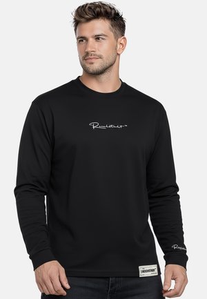 Mann trägt schwarzes Langarmshirt mit weißem Logo auf Brust, Bündchen und Saum, schaut leicht zur Seite vor einfarbigem Hintergrund.