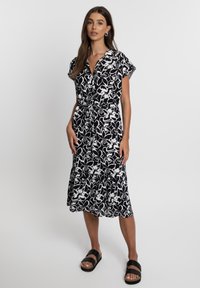 Schwarz-weiß gemustertes Kleid mit kurzen Ärmeln, Taillenknot, und gestuftem Saum, aus leichtem Stoff, kombiniert mit schwarzen Sandalen.