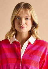 Červená a ružová pruhovaná polo tričko s bielym golierom, s placketom na gombíky a malým logom na hrudi. Mäkka textúra látky.