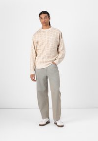 Beige Pullover mit einem strukturierten geometrischen Muster, kombiniert mit hellblauen geraden Jeans und weißen Sneakers. Einfaches Rundhalsausschnitt-Design.