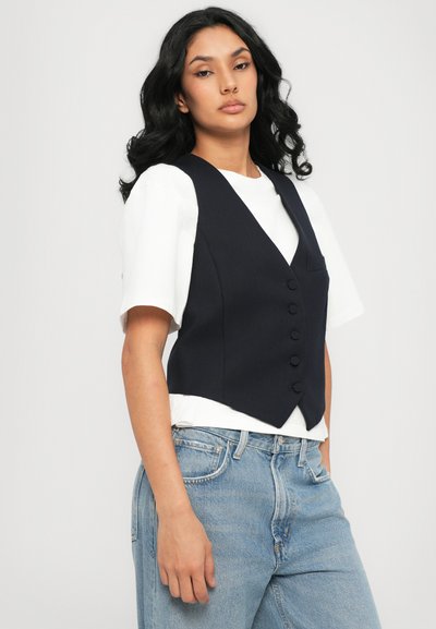 Gilets navy à cinq boutons, avec un ourlet pointu et une coupe slim, portés sur une chemise blanche. Jean en denim bleu clair avec une coupe décontractée.