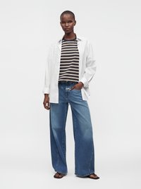 Mujer que lleva una camisa blanca abotonada sobre una camiseta de rayas, vaqueros azules de pierna ancha y sandalias marrones, de pie con una mano en el bolsillo.