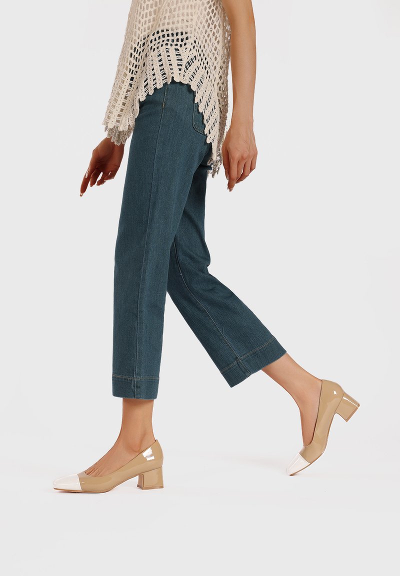 Décolleté in pelle lucida beige con punta bianca, abbinati a jeans cropped in denim azzurro chiaro e un top beige a maglia aperta con bordo a scallop.