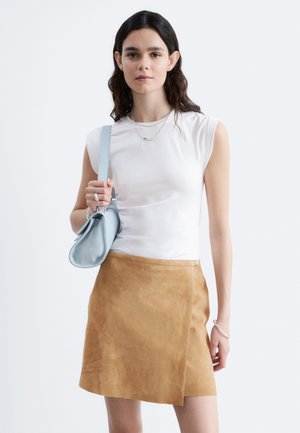 Femme aux longs cheveux foncés, portant un haut blanc sans manches, une jupe en daim beige, des bijoux en argent et tenant un sac à main bleu clair sur l'épaule.