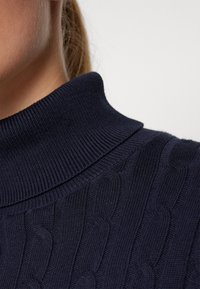 Maglione lavorato a maglia color navy con colletto alto a coste e motivo a treccia. La vista ravvicinata mette in evidenza la trama e i dettagli dei punti.