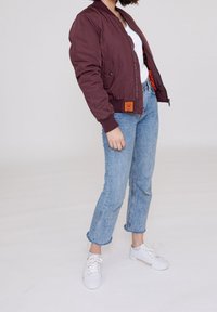 Giacca bomber bordeaux con polsini e orlo a costine. Fodera arancione visibile, abbinata a jeans azzurri chiari e sneaker bianche.