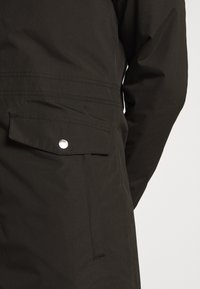Gros plan sur une manche de manteau noire et une poche latérale avec rabat et bouton-pression argenté sur fond blanc.