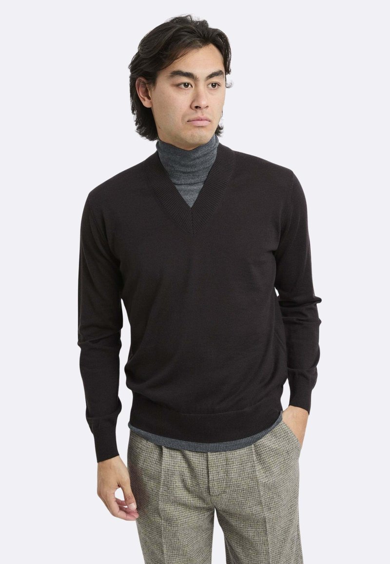 Uomo che indossa un maglione nero a scollo a V sopra un dolcevita grigio, con pantaloni beige a quadri, in piedi con una mano in tasca davanti a uno sfondo bianco.