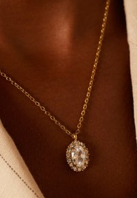 Gouden kettinkje met een ovale duidelijke edelsteen hanger, omringd door kleinere duidelijke stenen, weergegeven tegen een textuur van huid.