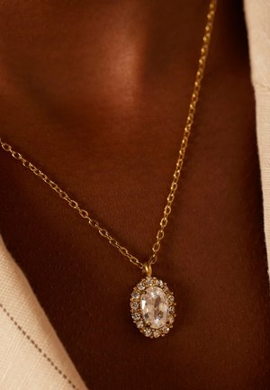 Collier en or avec un pendentif en pierre précieuse ovale et transparente, entouré de plus petites pierres claires, présenté sur un fond à texture de peau.
