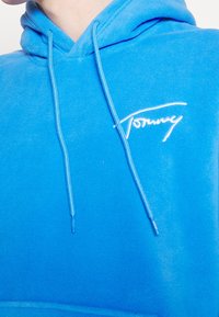 Felpa in pile blu con tasca frontale, cordini e logo "Tommy" ricamato in bianco. Tessuto morbido, vestibilità rilassata, colore vivace.