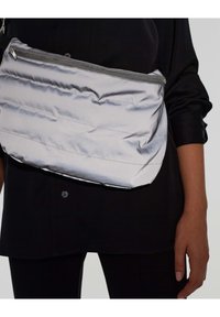 Borsa argentata a forma soffice con una texture liscia, caratterizzata da una forma curvata e una chiusura con zip. Indossata sotto il braccio, abbinata a una maglietta nera.