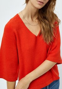 Maglione lavorato a maglia rosso, con scollatura a V e maniche corte, dal taglio rilassato. Texture morbida e design minimalista. Il modello indossa il capo con un paio di jeans casual.