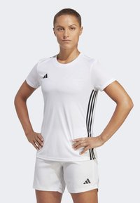 Bílé sportovní tričko s černým logem Adidas a třemi černými pruhy podél stran. Hladký materiál, krátké rukávy, v kombinaci s bílými šortkami.