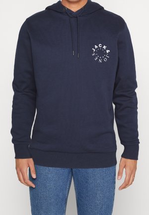 Homme portant un sweat à capuche bleu marine avec un logo circulaire blanc "Jack & Jones" sur la poitrine gauche, associé à un jean bleu.
