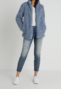 Vaaleansininen pörröinen takki, jossa on teksturoitu, kihara kangas. Päällä valkoinen paita, yhdistettynä kuluneisiin vaaleisiin farkkuihin ja valkoisiin tennareihin.