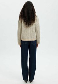 Nainen, jolla on pitkät kiharat hiukset, pukeutuneena beigeen ribbitakkiin, tummiin leveälahkeisiin muihin ja mustiin korkokenkiin, seisoo selkä kameraan.