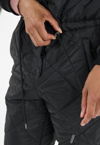 Zwarte geblokte jas met trekkoorden; heeft een rits, een gladde textuur en contrasterende zwarte accenten. Handen die het trekkoord aanpassen.