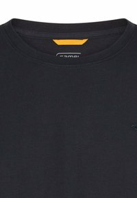 Close-up van een zwart T-shirt met ronde hals, met een klein geel stoffen label aan de binnenkant van de kraag en vage logo-stiksels op de rechterborst.