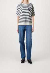 Person som bär en marinblå och vit randig kortärmad skjorta, blå raka jeans och svarta skor, stående mot en neutral vit bakgrund.