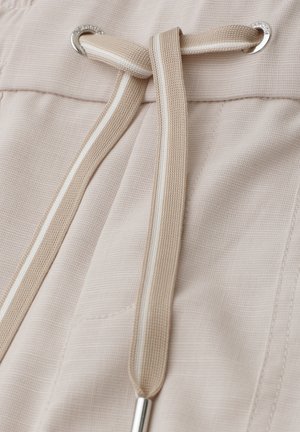 Gros plan d’un pantalon en tissu beige avec un cordon assorti passé dans des œillets métalliques, noué en un nœud simple.