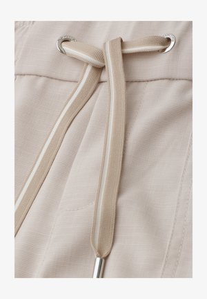 Close-up of beige fabric trousers with a matching drawstring threaded through metal eyelets, tied in a simple knot.
