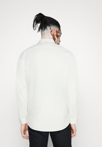 Camisa blanca de manga larga con un diseño limpio, puños abotonados y cuello. La tela parece suave y ligera. Vista desde la parte de atrás.