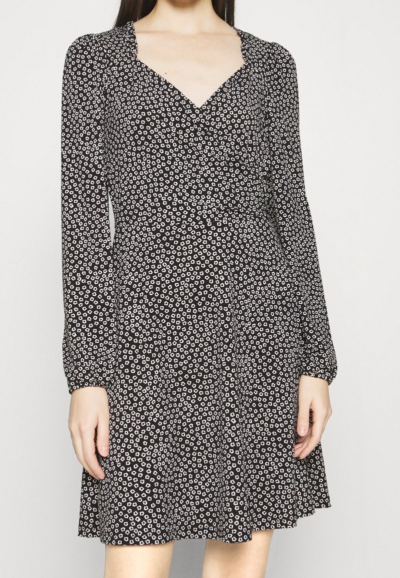 Femme portant une robe noire à manches longues avec un petit imprimé floral blanc et un décolleté en V, debout devant un fond blanc uni.