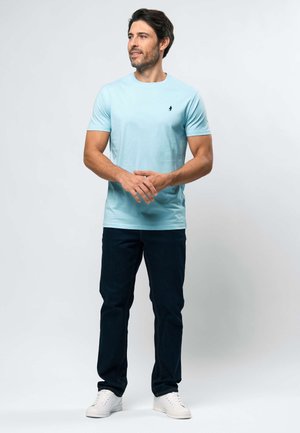 Muž stojaci s rukami zloženými, oblečený v svetlomodrom tričku, tmavých džínsoch a bielych teniskách, pozerajúci sa doľava.