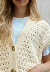 Gilet en tricot crème avec un motif en maille ouverte texturé, doté d'un col en V, de manches courtes et de grands boutons marron sur une chemise bleu clair.