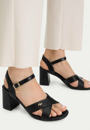 Fødder iført sorte højhælede sandaler med ankelremme og gyldne logocharms, kombineret med off-white vidde bukser.