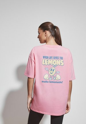 Frau mit braunem Pferdeschwanz trägt ein pinkfarbenes T-Shirt mit einem Comic-Zitronenmotiv und dem Text "Wenn dir das Leben Zitronen gibt, mach Limonade daraus" auf der Rückseite.